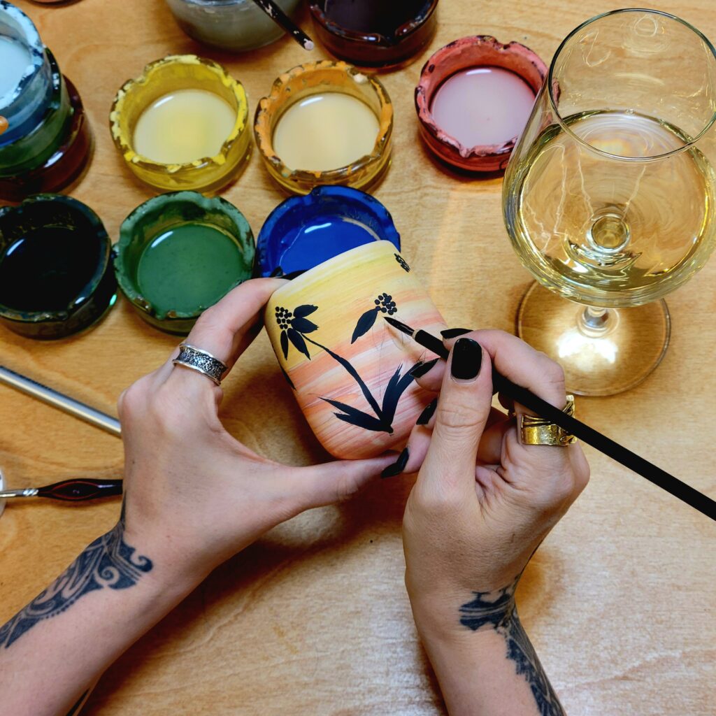 Close-up of an artist's hands carefully painting black floral illustrations onto a sunset-colored ceramic tumbler in the Corax Ceramics studio.