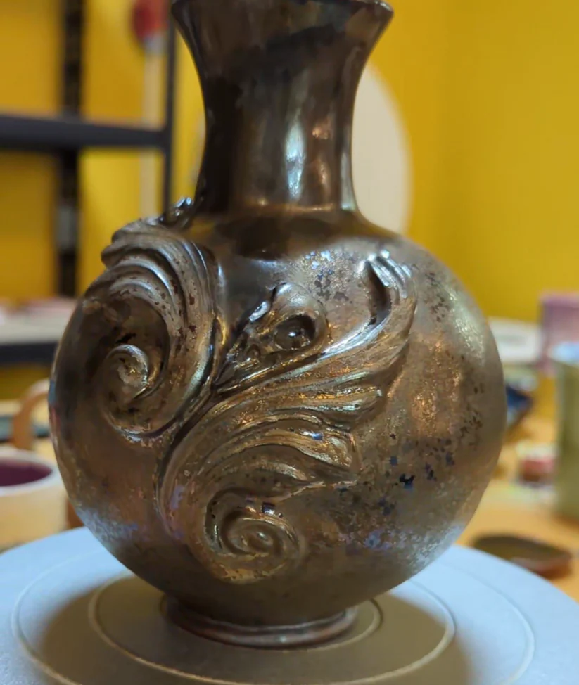 Handmade gothic stoneware vase with metallic bronze glaze and intricate relief carvings, held by a ceramicist.
